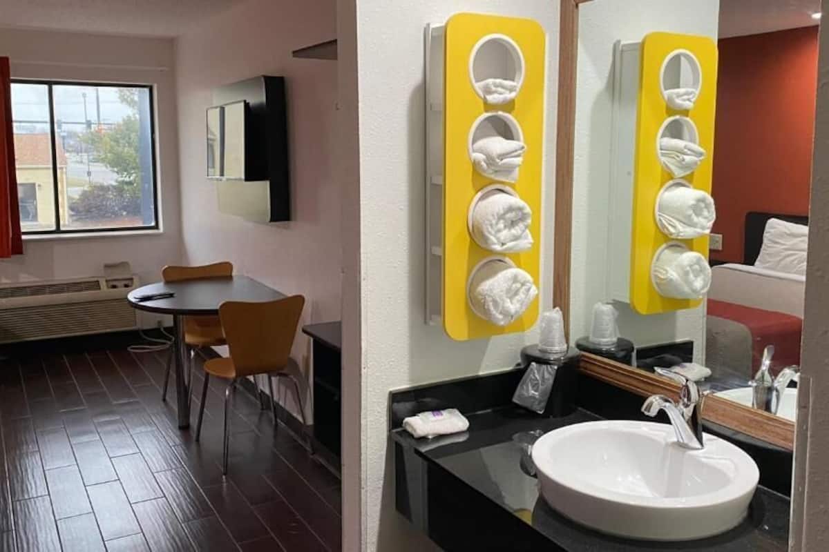 A hotel room with a dining area, a large window with curtains, a round table, and a modern bathroom with a sink and mirror.
