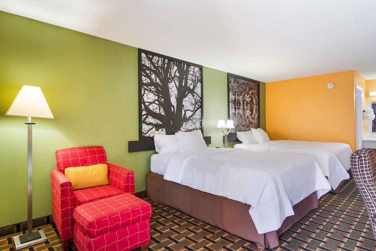 A hotel room with a bed, two bedside tables, a red armchair, a floor lamp, and a large tree artwork on the wall.