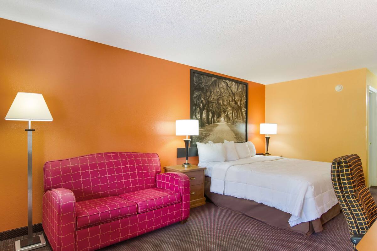 A hotel room with a red sofa, a bed with white bedding, two bedside lamps, and a painting on the wall.
