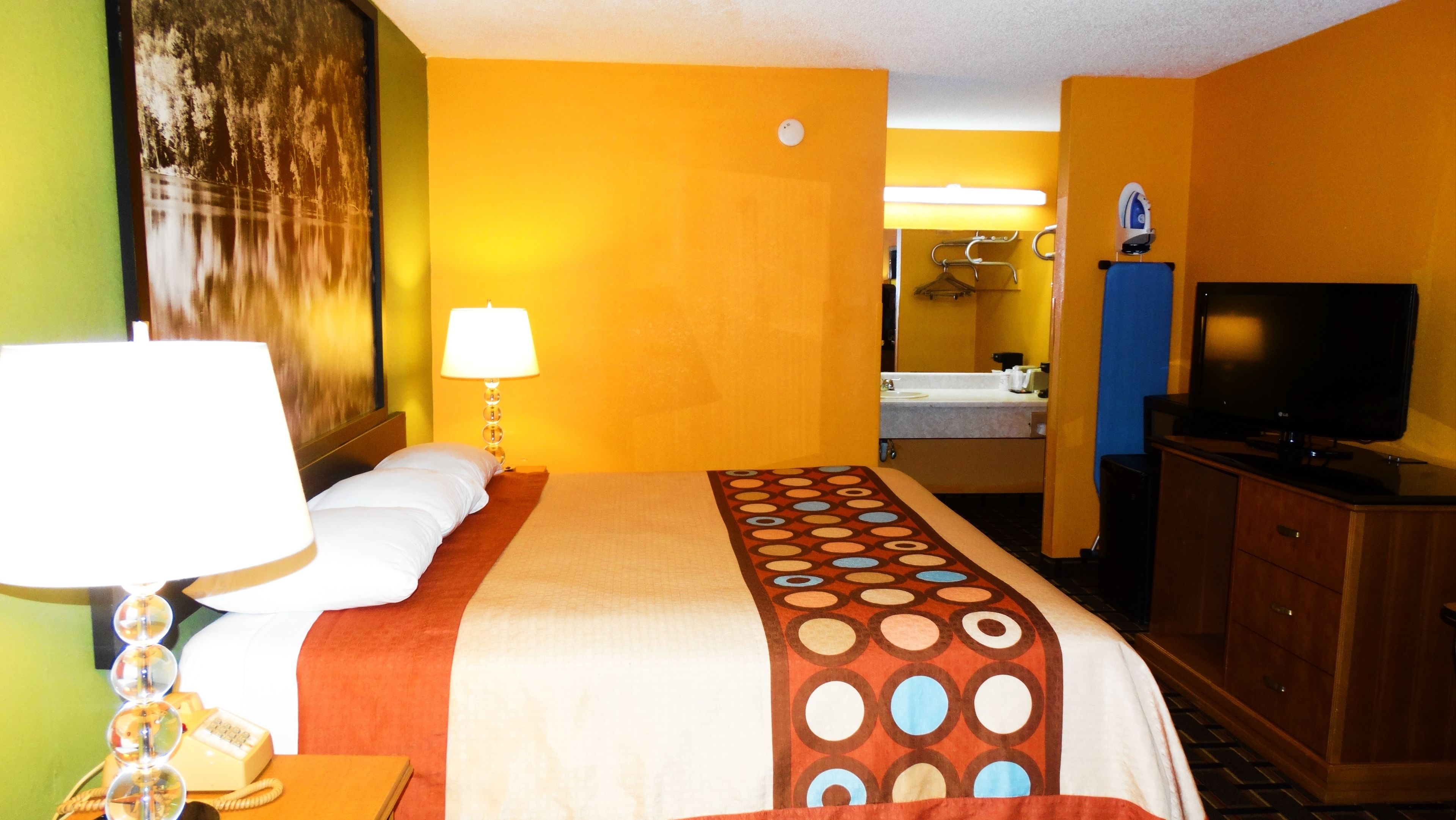Habitación de hotel con cama, televisor, lámpara, espejo y baño visible a través de una puerta abierta.