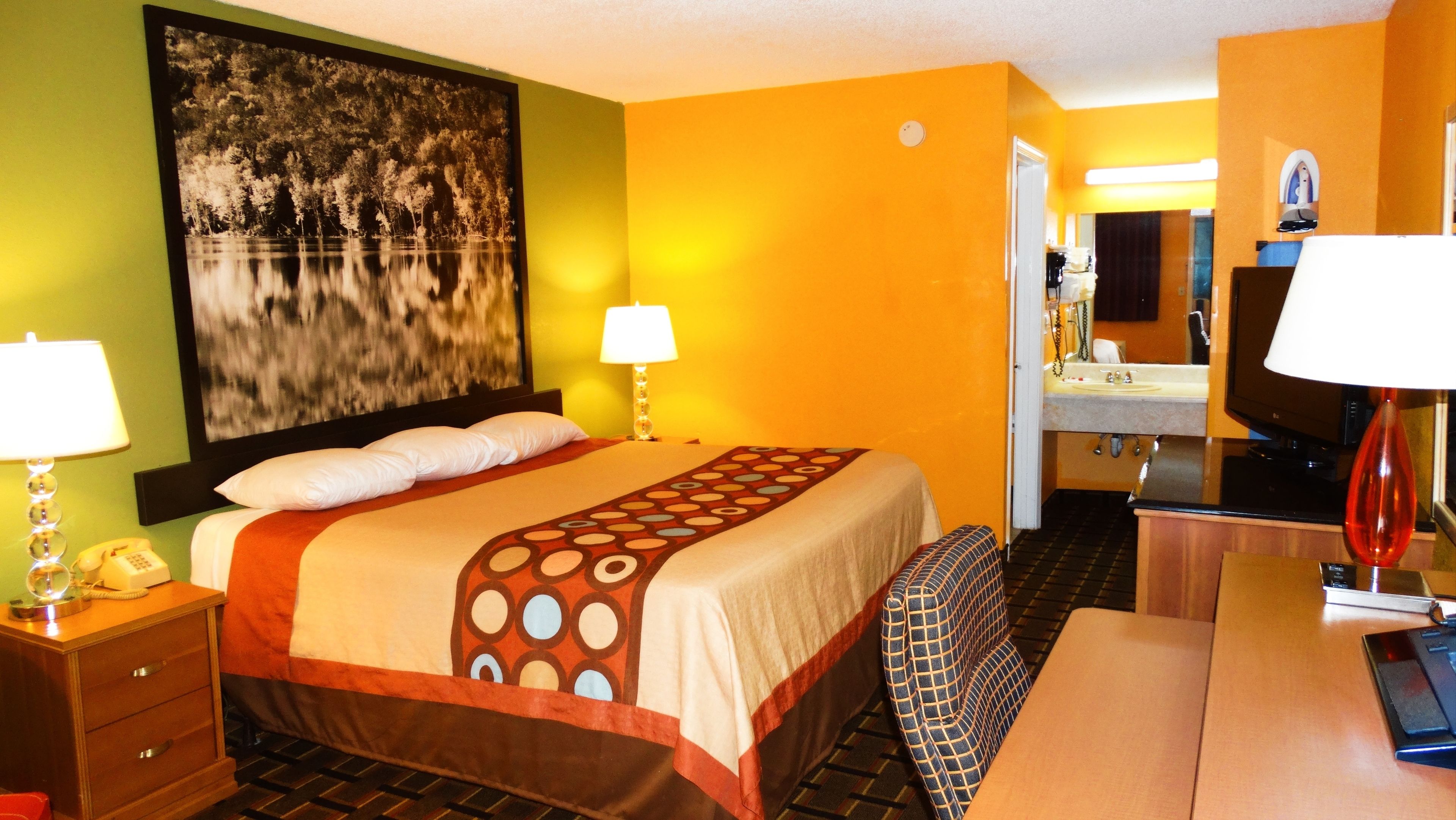 A hotel room with a bed, bedside lamps, a desk with a computer, a television, and a bathroom visible through an open door.