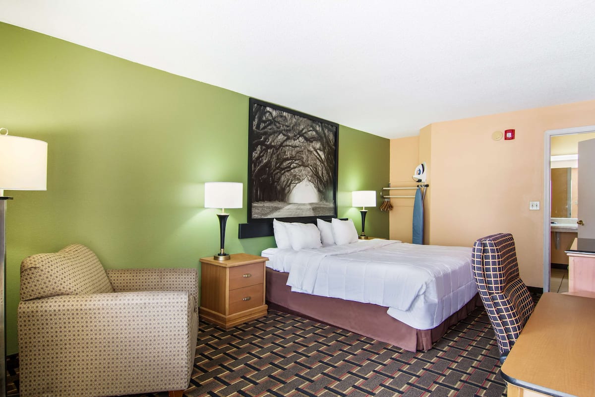 A hotel room with a bed, a chair, a desk, and a painting on the wall.
