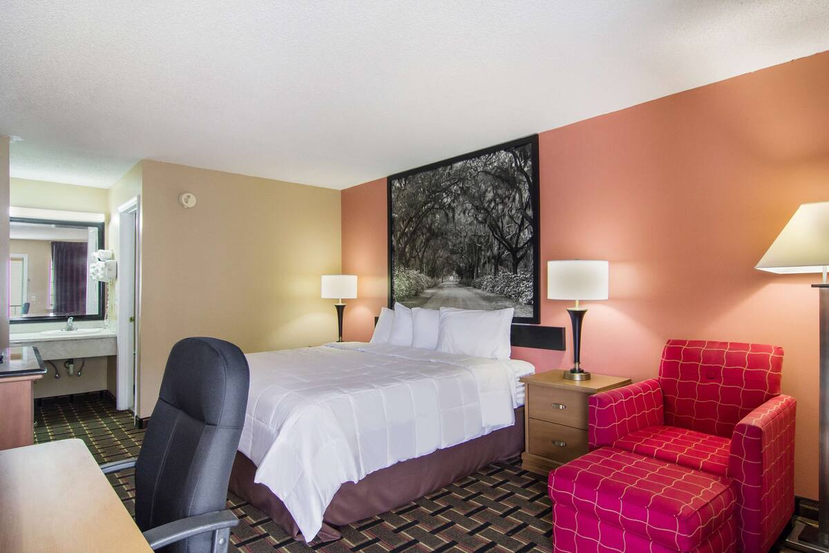 A hotel room with a bed, a desk, a chair, a nightstand, and a painting on the wall.