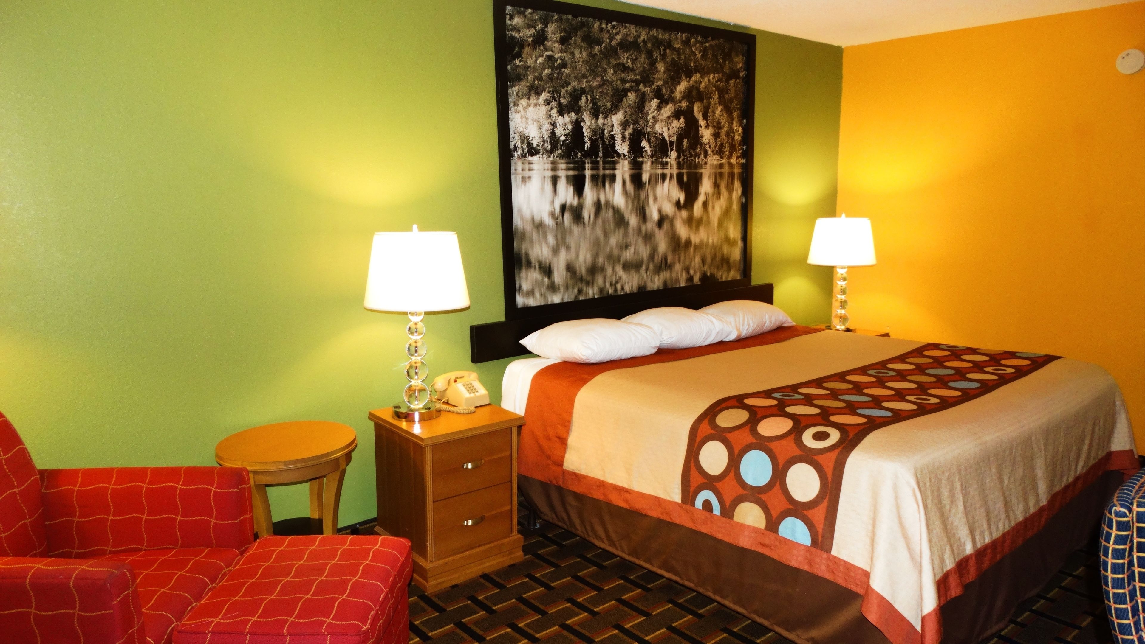 Ein Hotelzimmer mit einem Bett, zwei Nachttischlampen, einem Telefon, einem Sessel und einem Bild an der Wand.