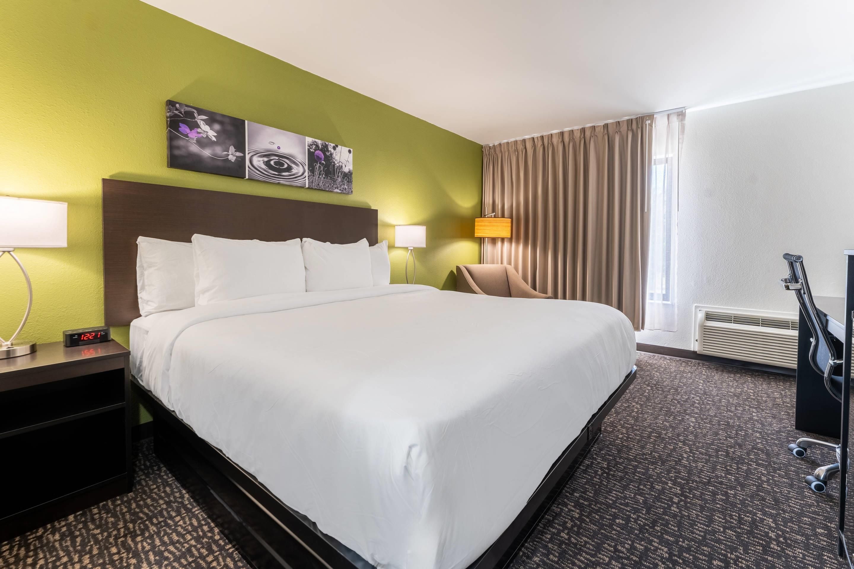 A hotel room with a large bed, two bedside tables, a chair, a desk, a lamp, a painting, and a window with curtains.