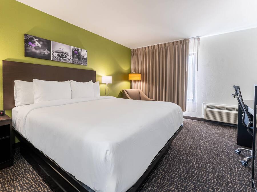 A hotel room with a large bed, two bedside tables, a chair, a desk, a lamp, a painting, and a window with curtains.