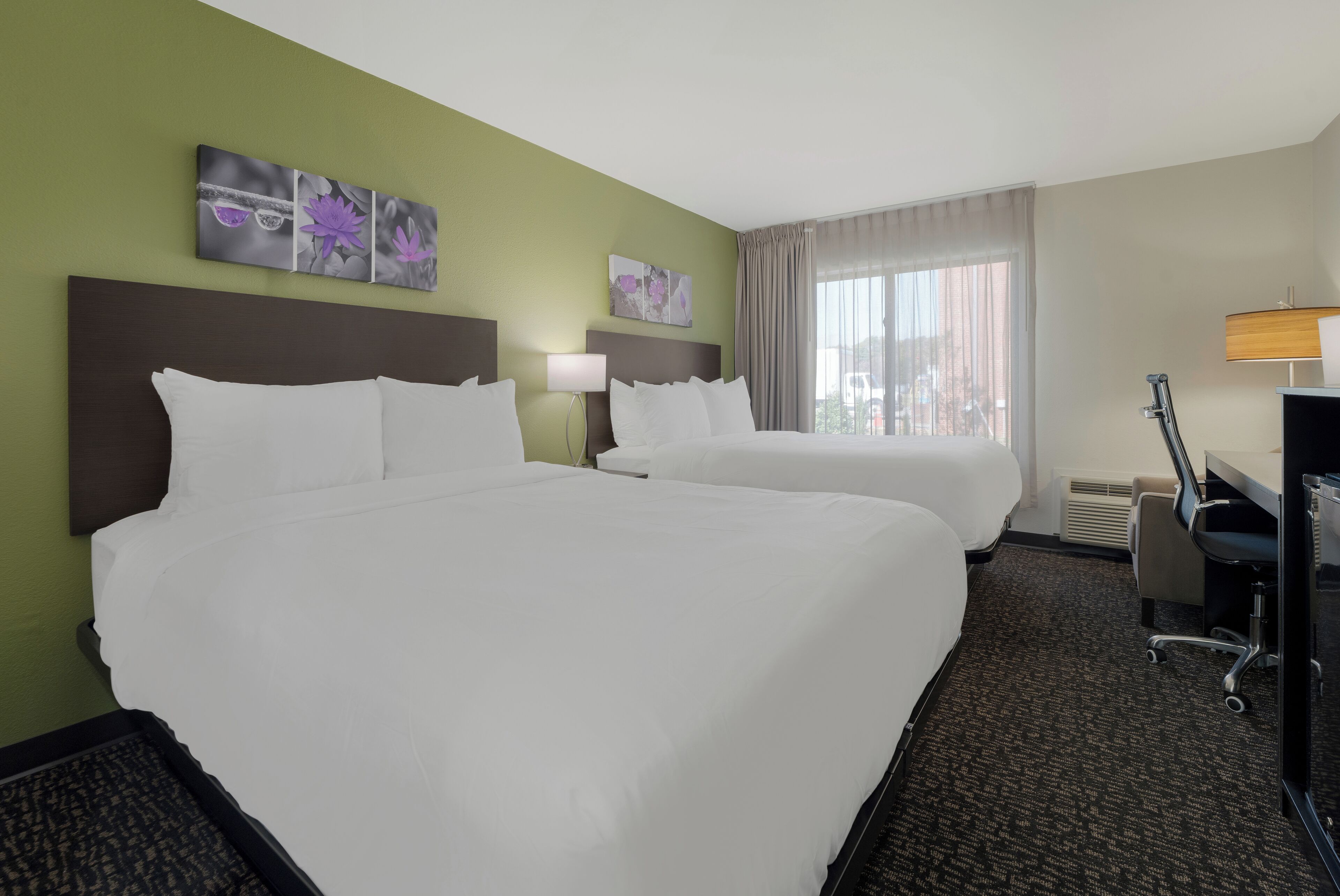 A hotel room with two beds, a desk, a chair, a lamp, and artwork on the walls.