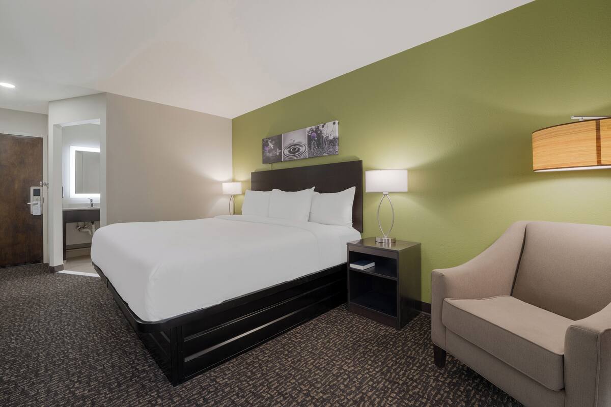 A hotel room with a large bed, a nightstand with a lamp, a chair, and a green accent wall.
