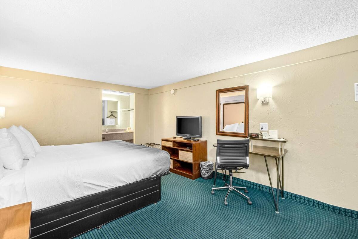A hotel room with a bed, a desk with a television, a chair, and a small table with a lamp.
