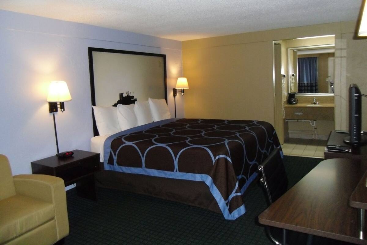 A hotel room with a bed, two bedside lamps, a desk, a chair, and a bathroom visible through an open door.
