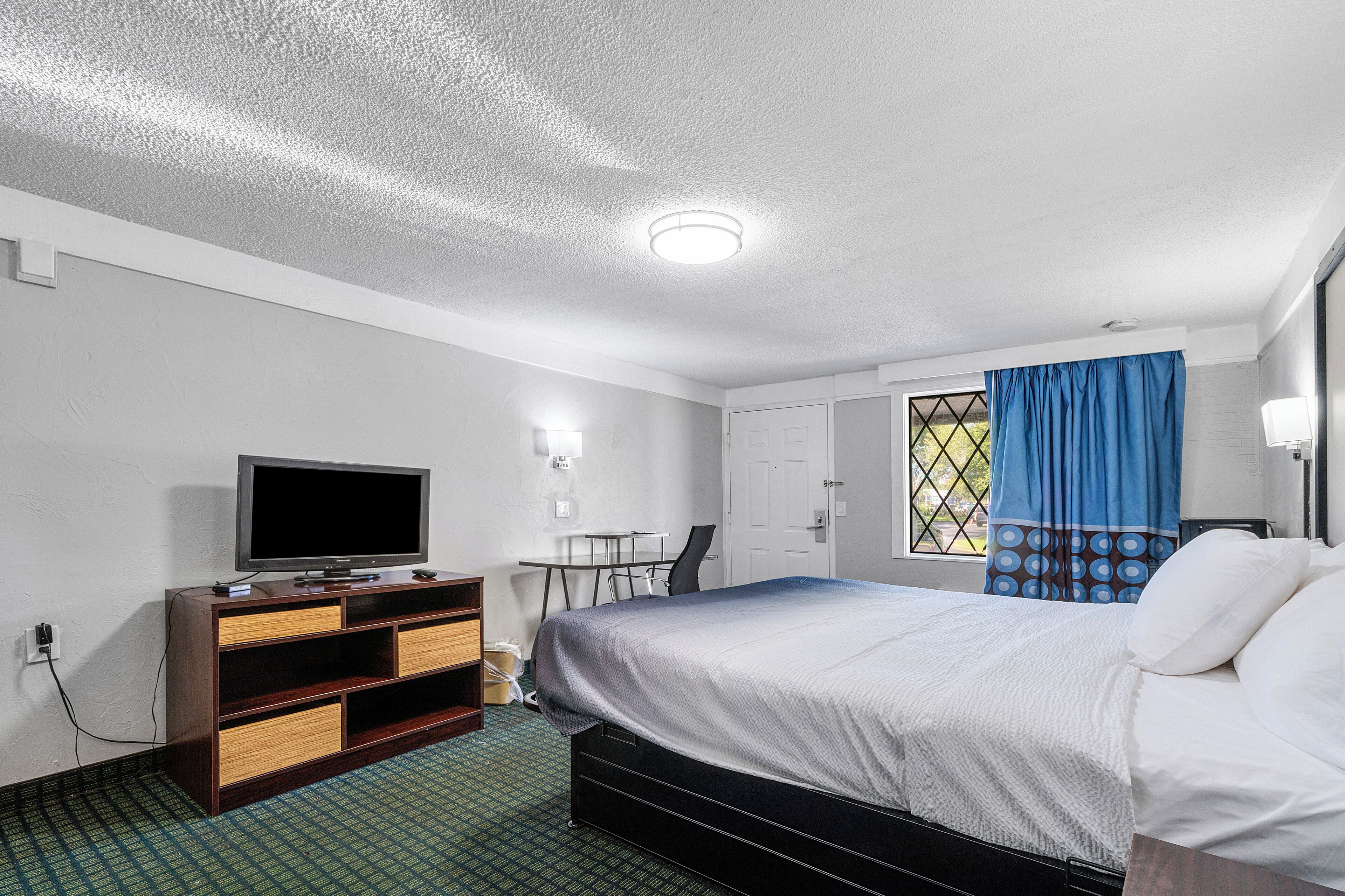 A hotel room with a bed, a TV on a wooden dresser, a desk with a chair, and a window with blue curtains.