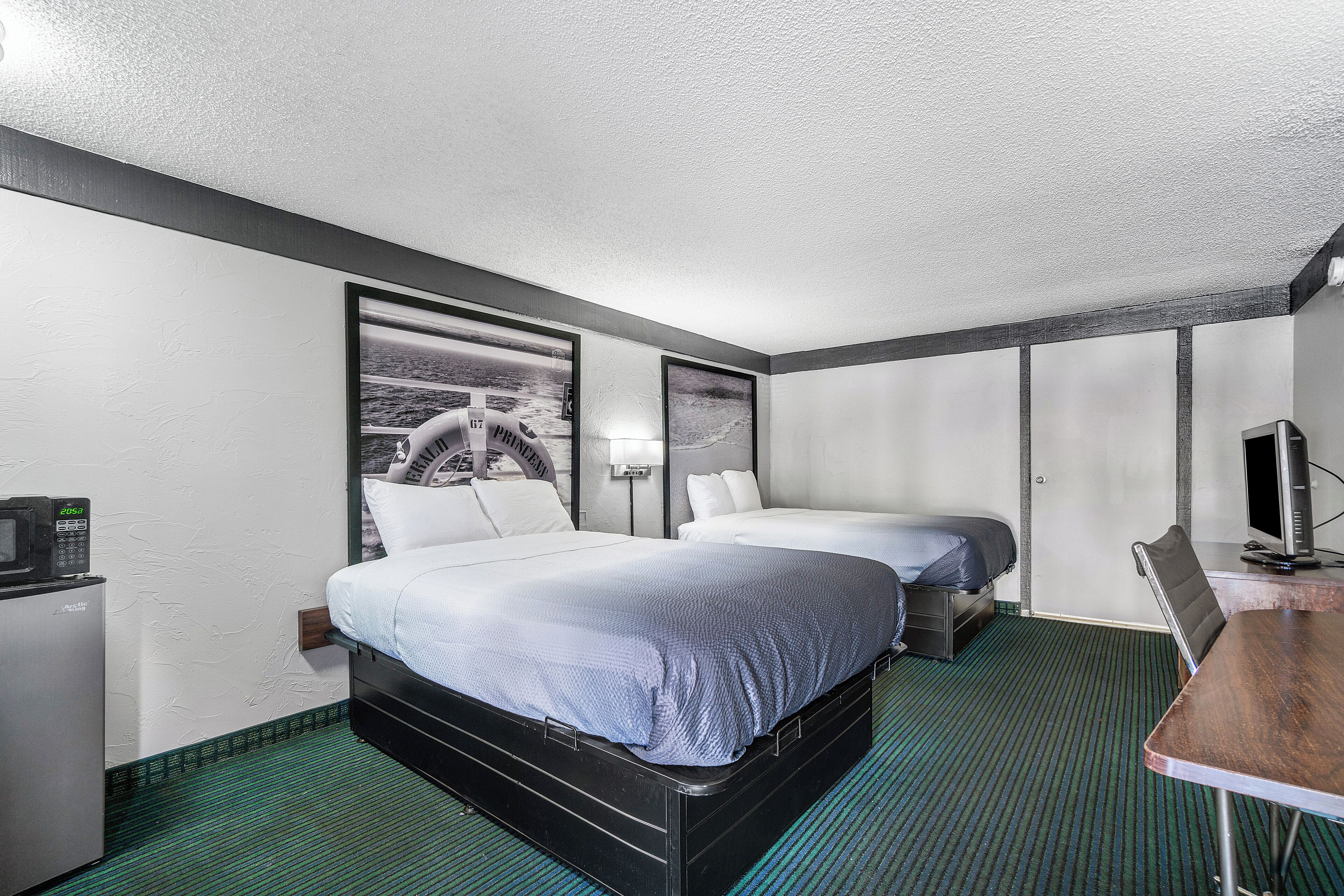 Habitación de hotel con dos camas, un escritorio, una silla, un televisor, un refrigerador y una lámpara fijada a la pared.
