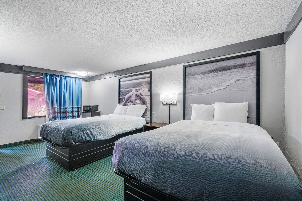 Une chambre d’hôtel avec deux lits, une grande fenêtre avec des rideaux bleus et des œuvres d’art accrochées aux murs.