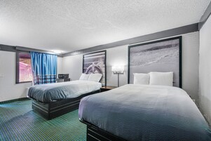 A hotel room with two beds, a large window with blue curtains, and artwork on the walls.