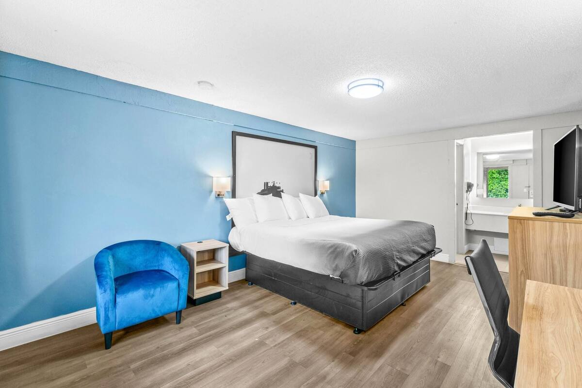 A modern hotel room with a large bed, a blue armchair, a wooden desk, and a mirror.