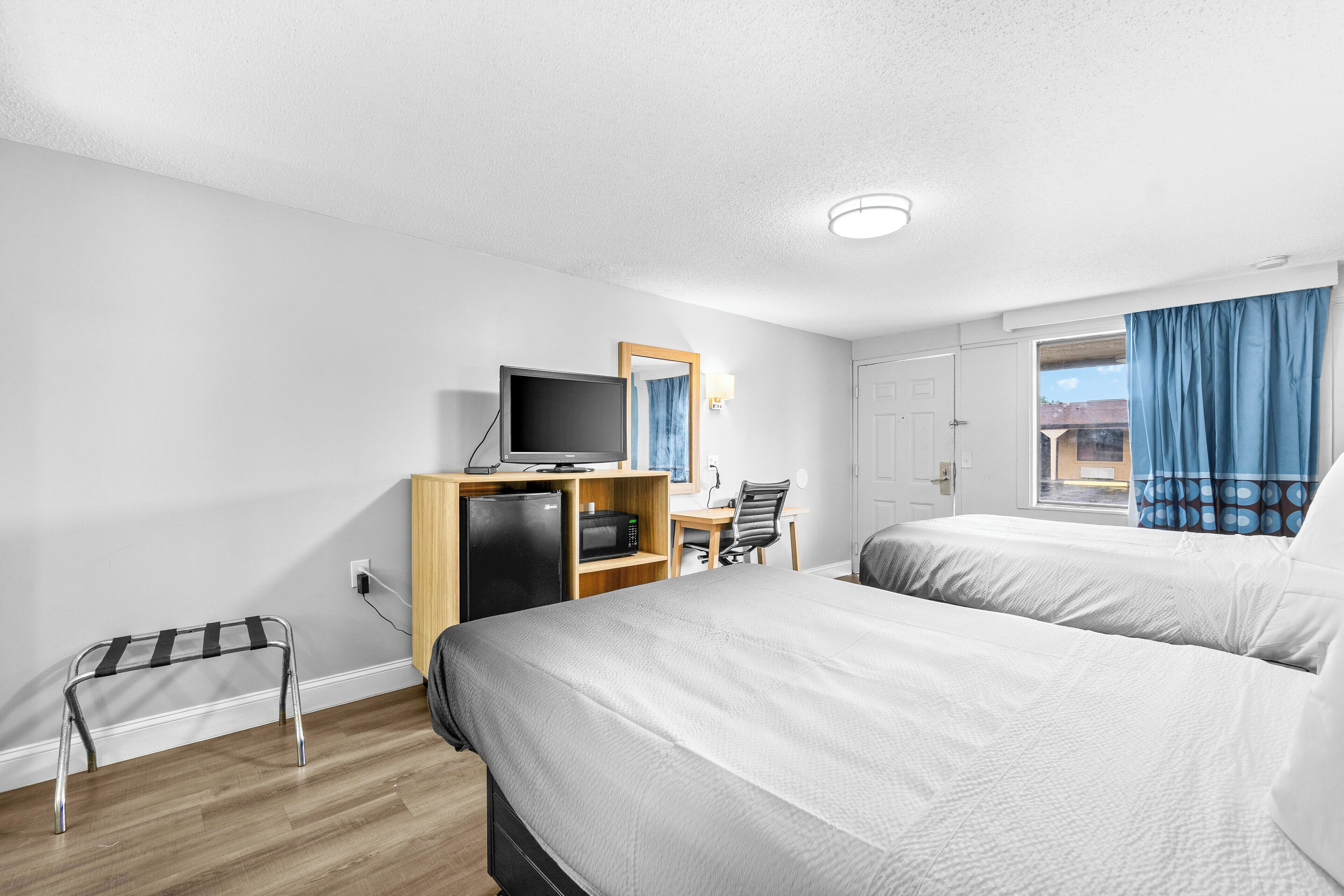 Una habitación de hotel con dos camas, un escritorio con computadora, un televisor y un refrigerador pequeño.