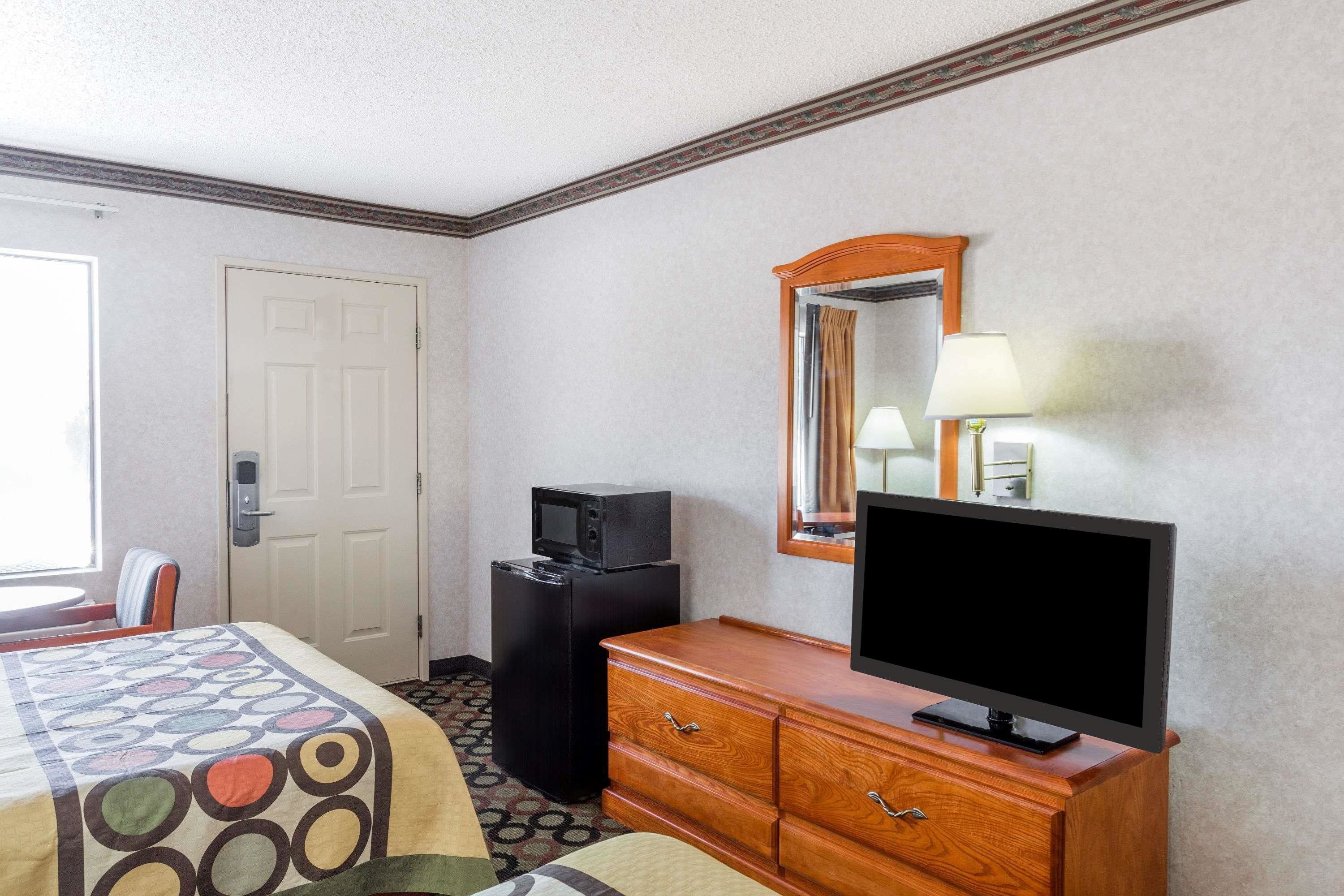 A hotel room with a bed, a television on a wooden dresser, a small black refrigerator, and a mirror on the wall.