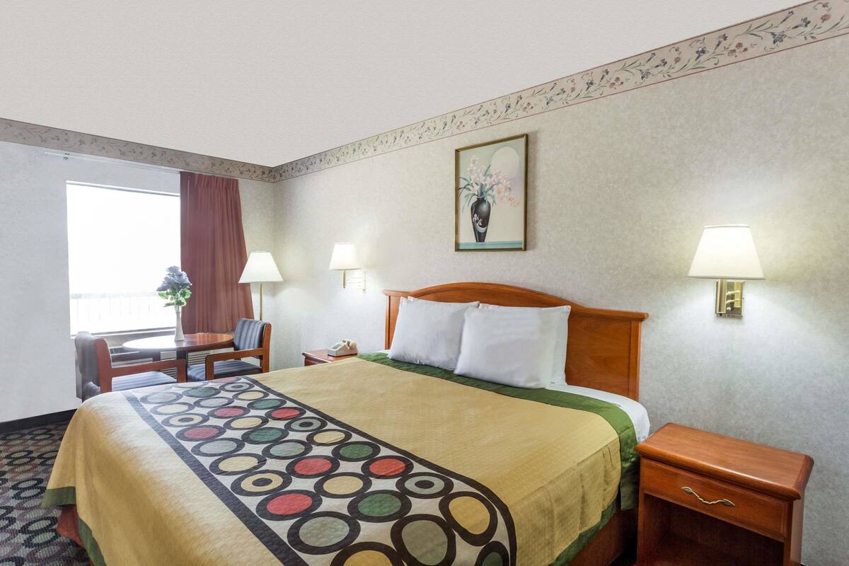 Quarto de hotel com uma cama grande, duas luminárias de cabeceira, uma mesa de cabeceira de madeira, uma escrivaninha com cadeira e uma janela com cortinas.