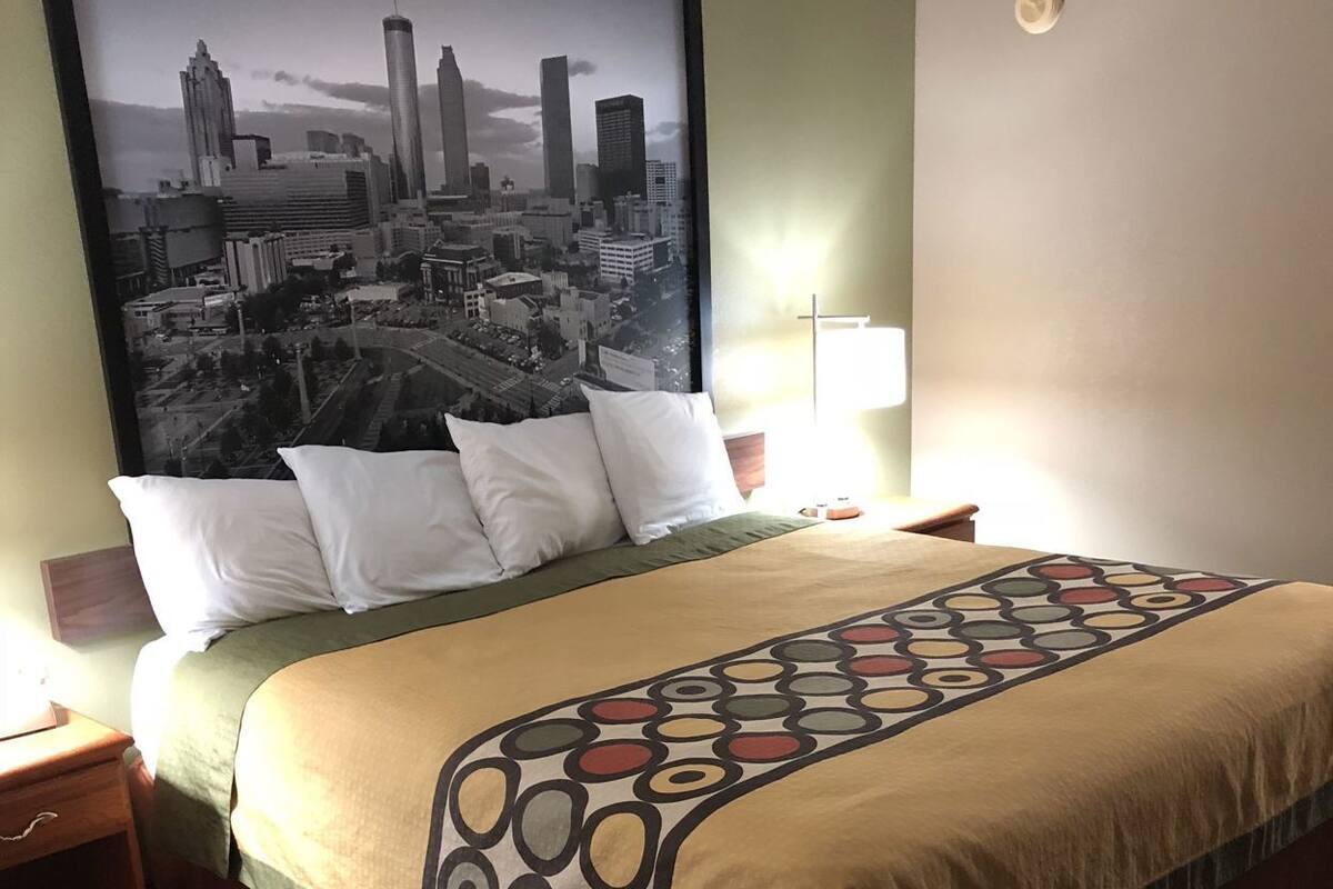 Quarto de hotel com uma cama, um pôster de paisagem urbana, uma mesinha de cabeceira, um abajur e um tapete estampado.