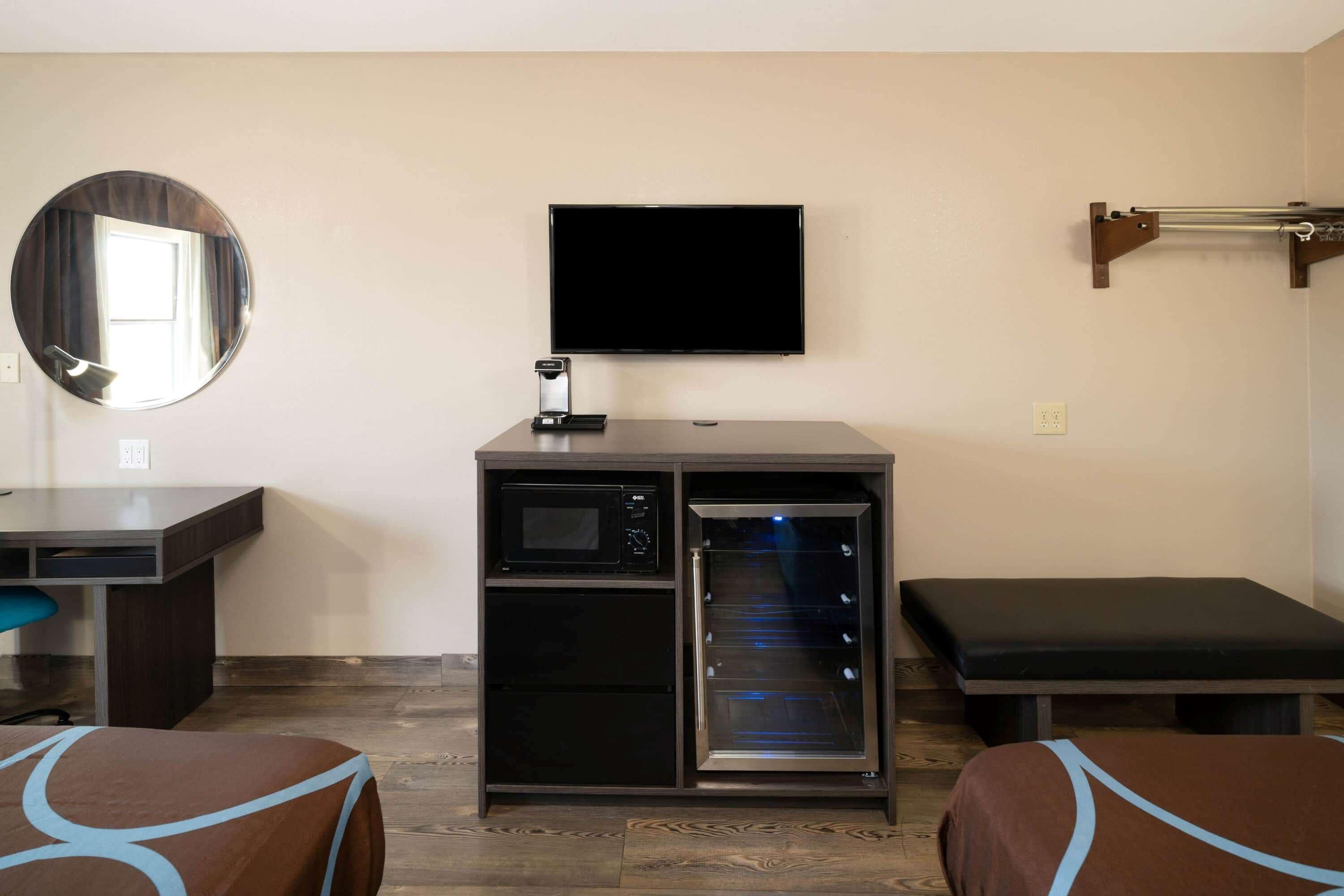 A compact room with a flat-screen TV, a microwave, a mini-fridge, a bench, and a bed.