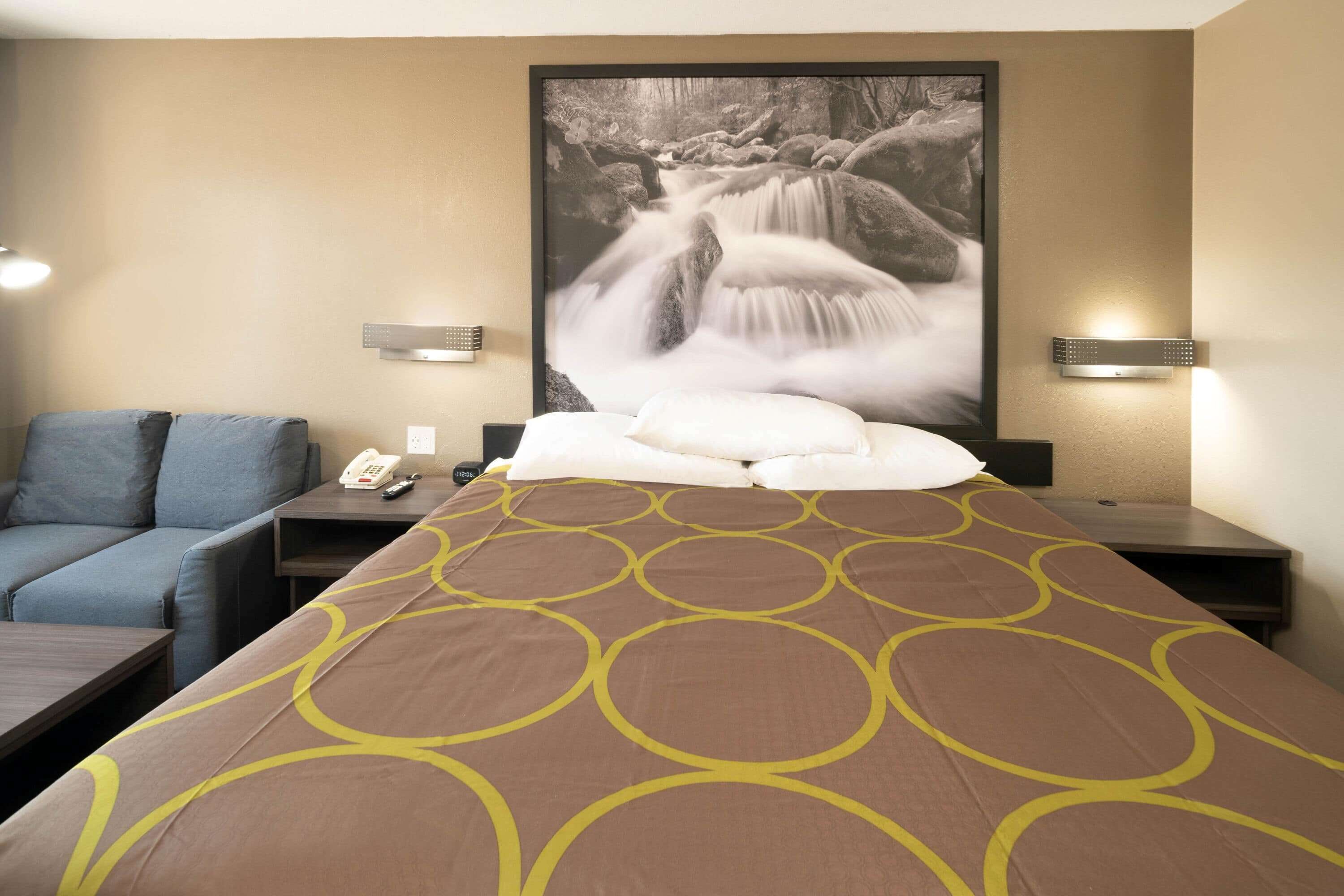 A hotel room with a bed, a sofa, a nightstand with a phone and remote, and a wall-mounted light fixture.