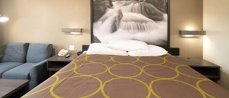 A hotel room with a bed, a sofa, a nightstand with a phone and remote, and a wall-mounted light fixture.