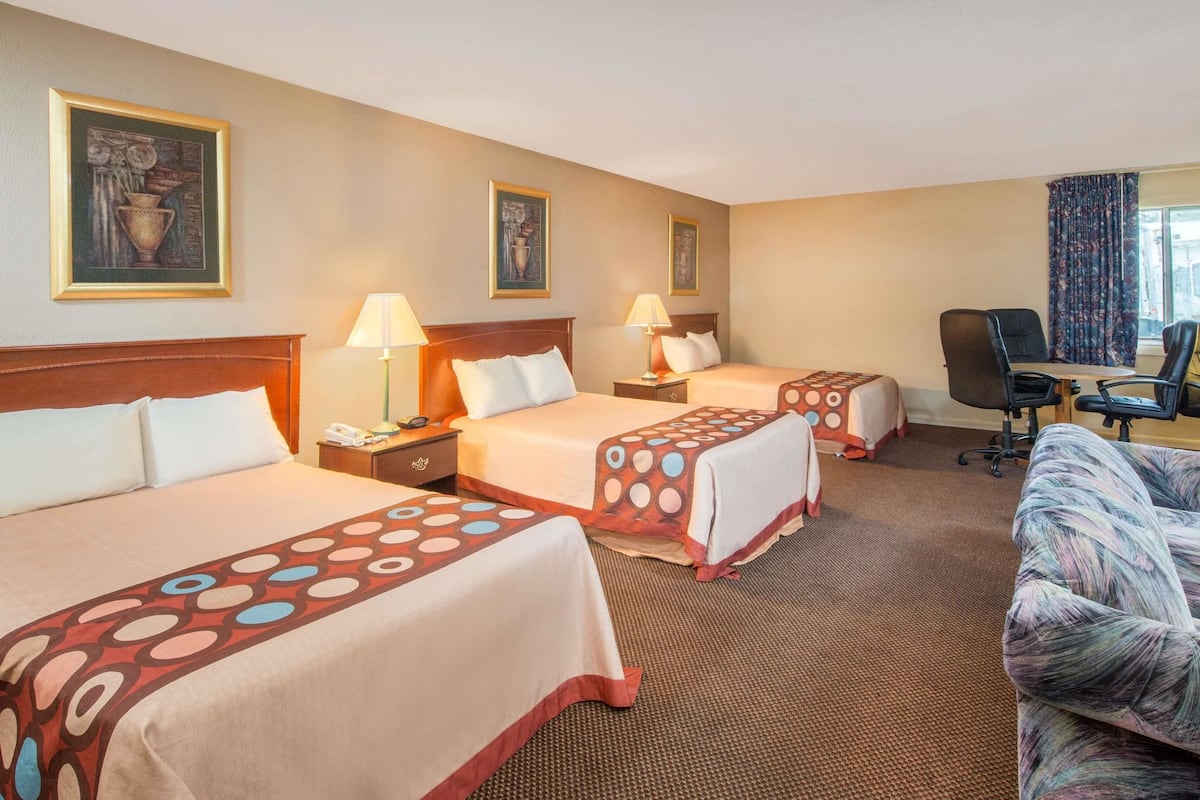 Quarto de hotel com três camas, uma escrivaninha e uma cadeira. Há quadros nas paredes e uma janela com cortinas.