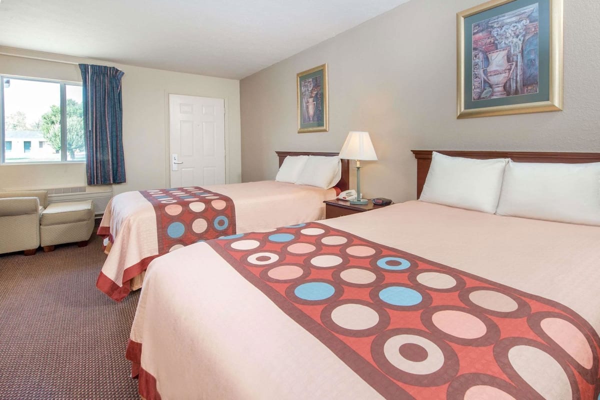 Quarto de hotel com duas camas, uma escrivaninha, uma cadeira, um abajur e um quadro na parede.
