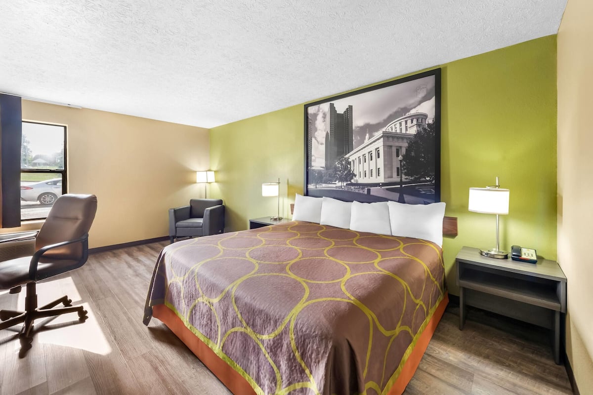 Een hotelkamer met een groot bed, twee nachtkastjes, een bureau met stoel en een raam met uitzicht op een gebouw en een auto.