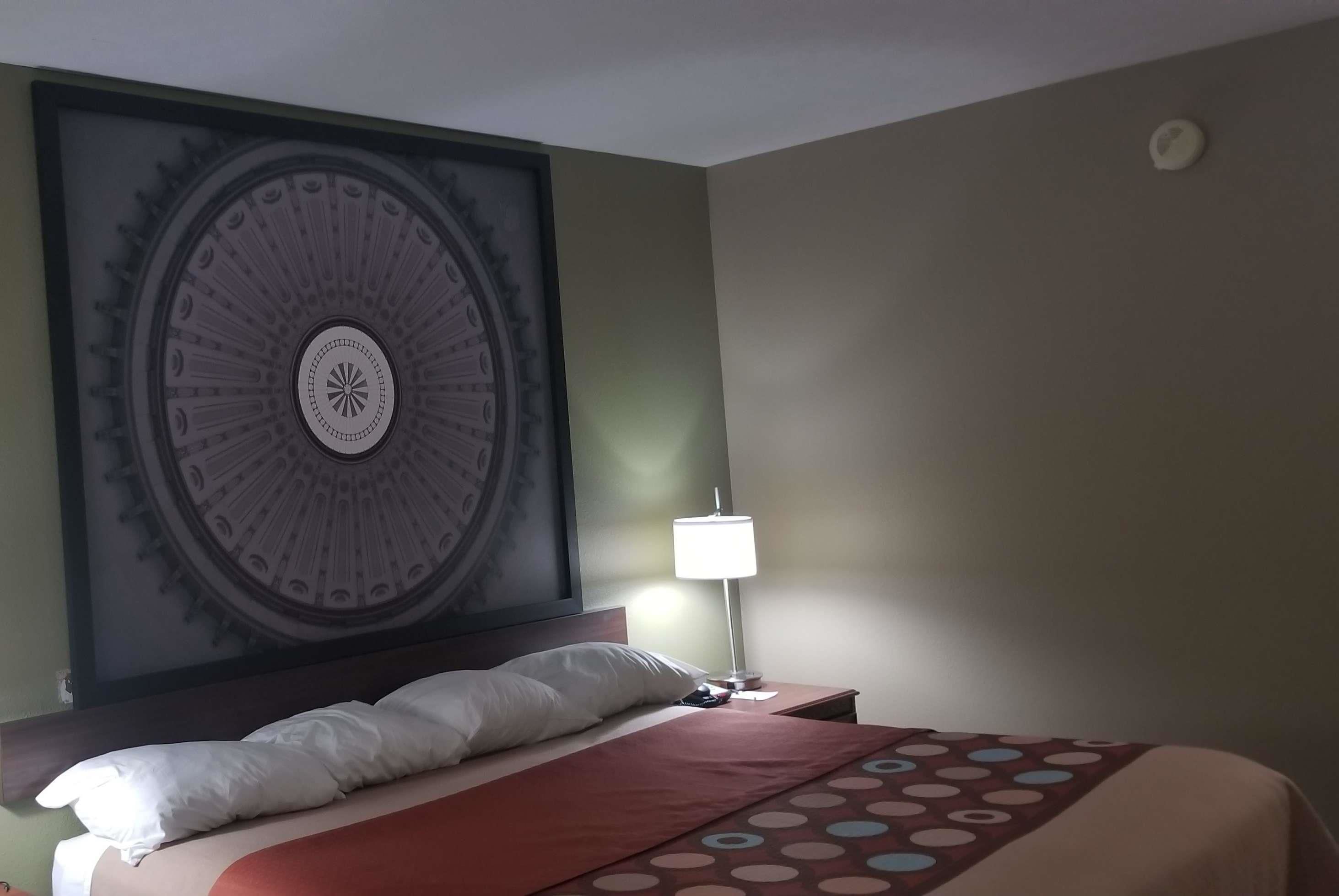Habitación de hotel con un gran adorno circular en la pared, una cama con colcha rojo y beige, un almohadón blanco y una mesita de noche con lámpara.