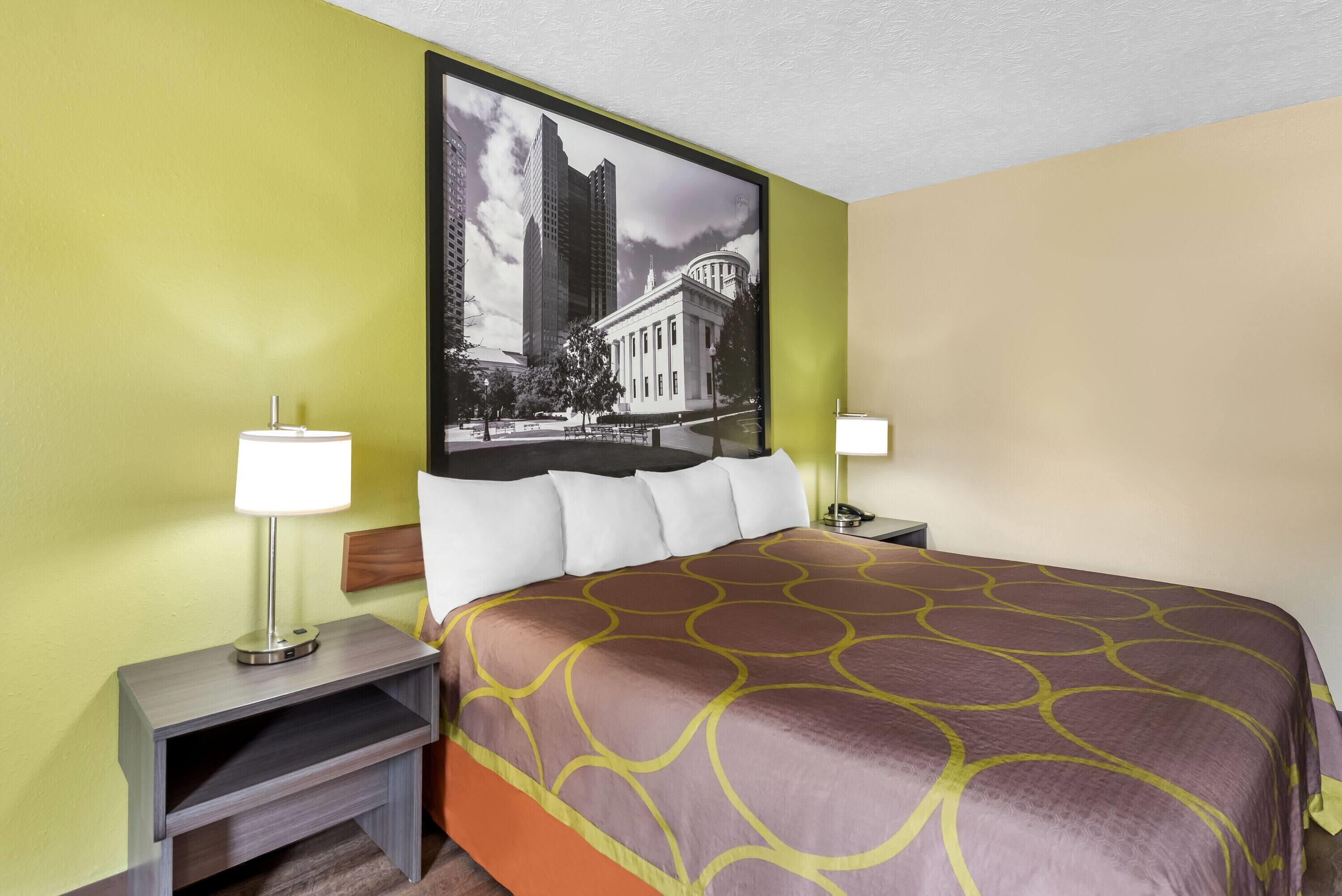 Habitación de hotel con una cama grande, dos mesitas de noche con lámparas, un televisor montado en la pared y un cuadro con una vista urbana.