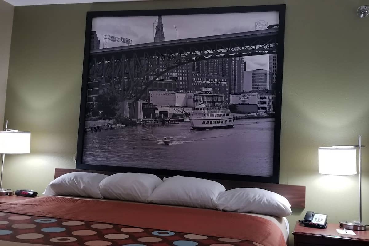 Een hotelkamer met een groot gepoft schilderij van een brug, een bed met witte kussens, twee nachtlampjes en een bureau met een telefoon en een lamp.
