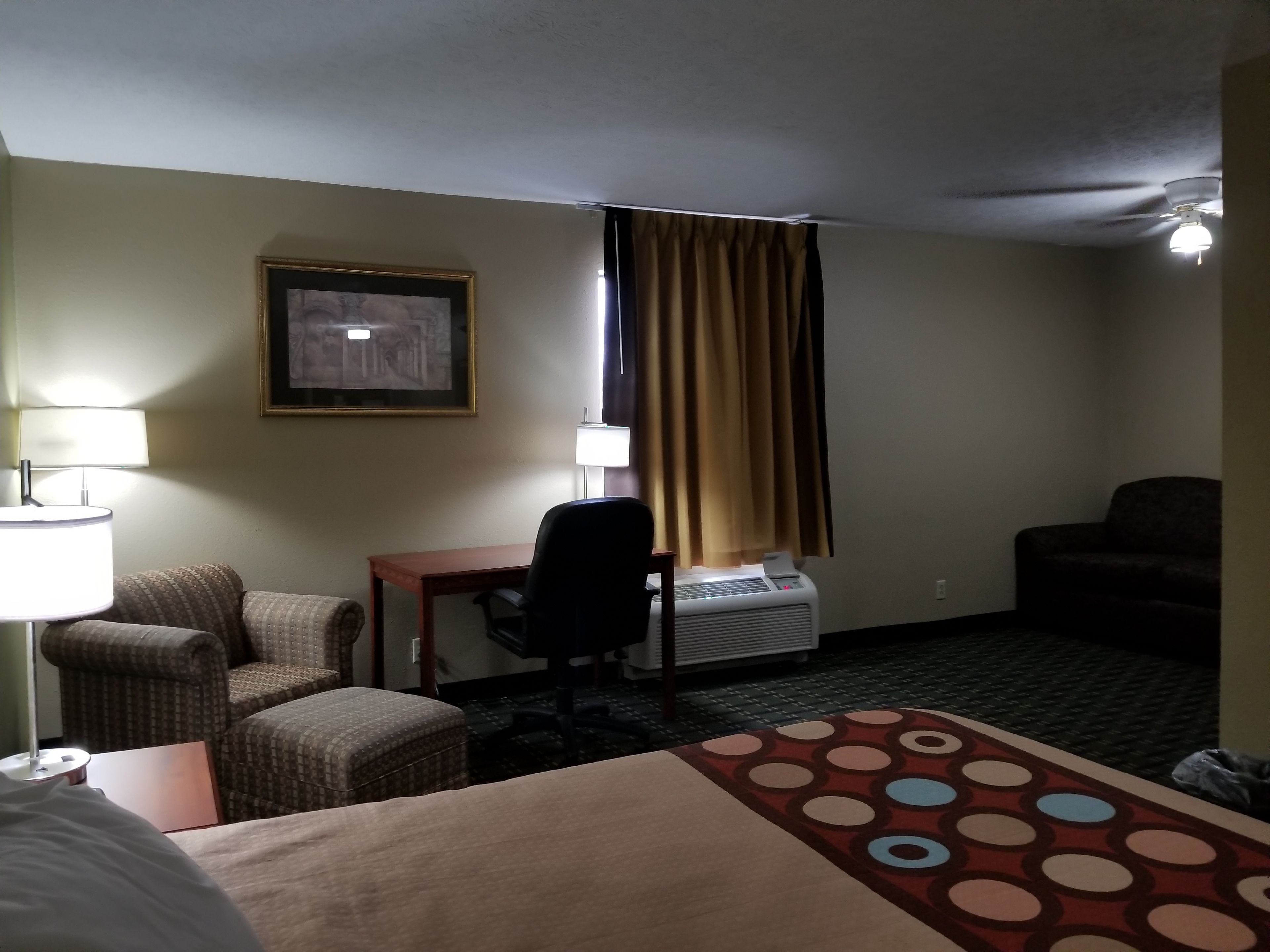 Ein Hotelzimmer mit einem Bett, einem Schreibtisch, einem Stuhl, einer Lampe, einem Heizkörper und einem Deckenventilator.