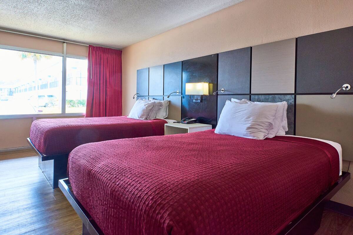 Ein Hotelzimmer mit zwei Betten, einem Nachttisch mit Lampe und einem Fenster mit Blick nach draußen.