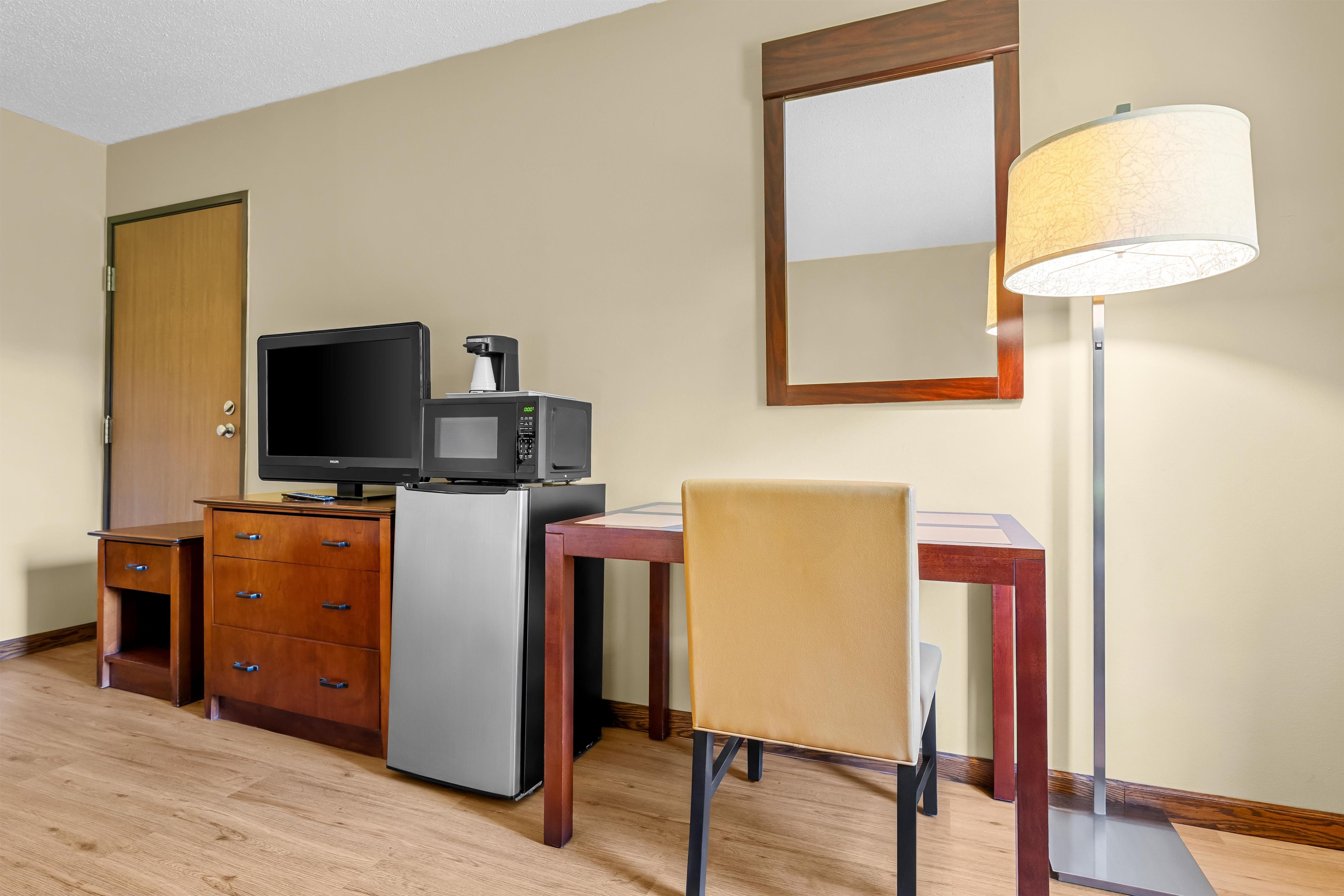 A compact hotel room with a kitchenette, a flat-screen TV, a refrigerator, a wooden desk, a chair, a floor lamp, and a mirror.