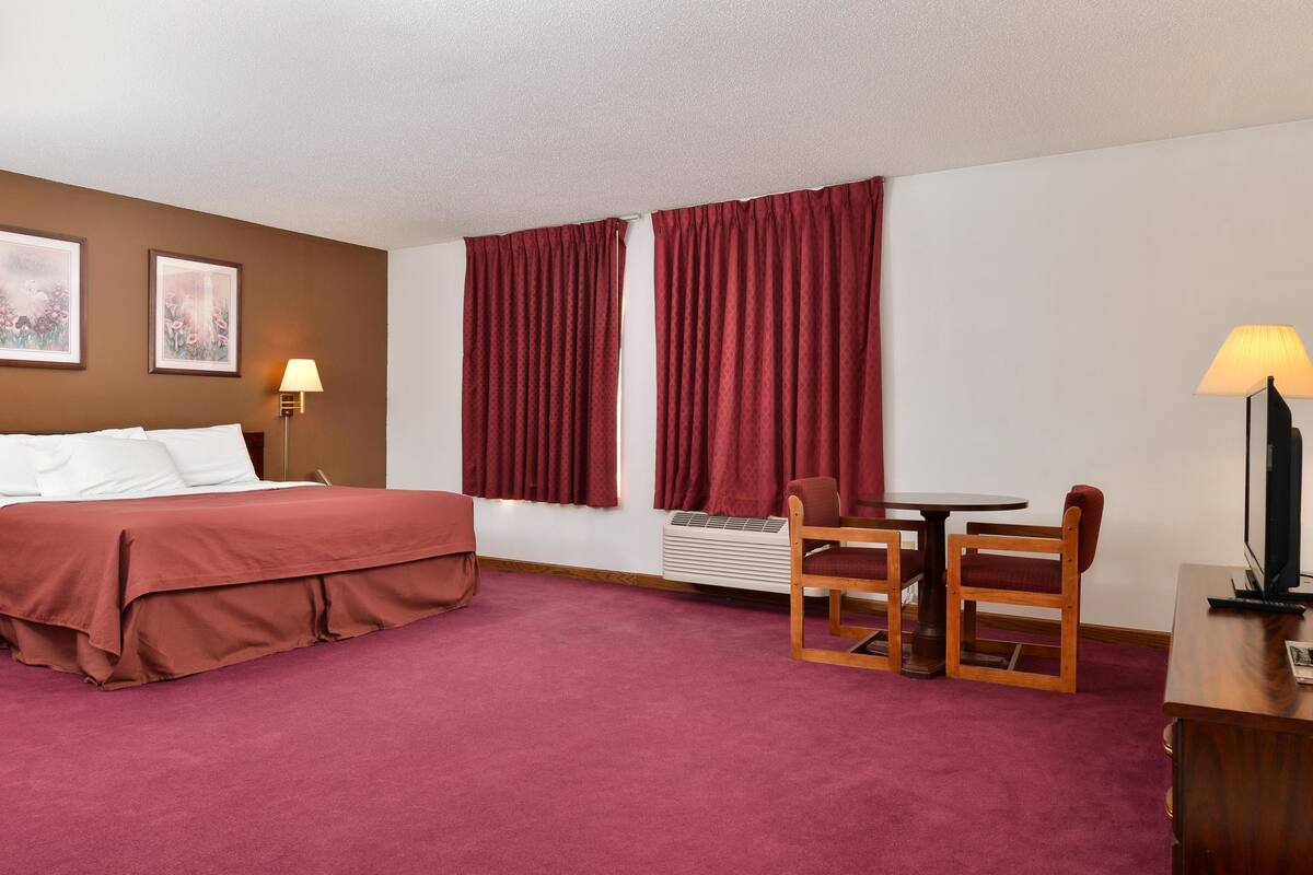 Une chambre d’hôtel avec un lit, un bureau, une chaise, une lampe et une fenêtre avec des rideaux.