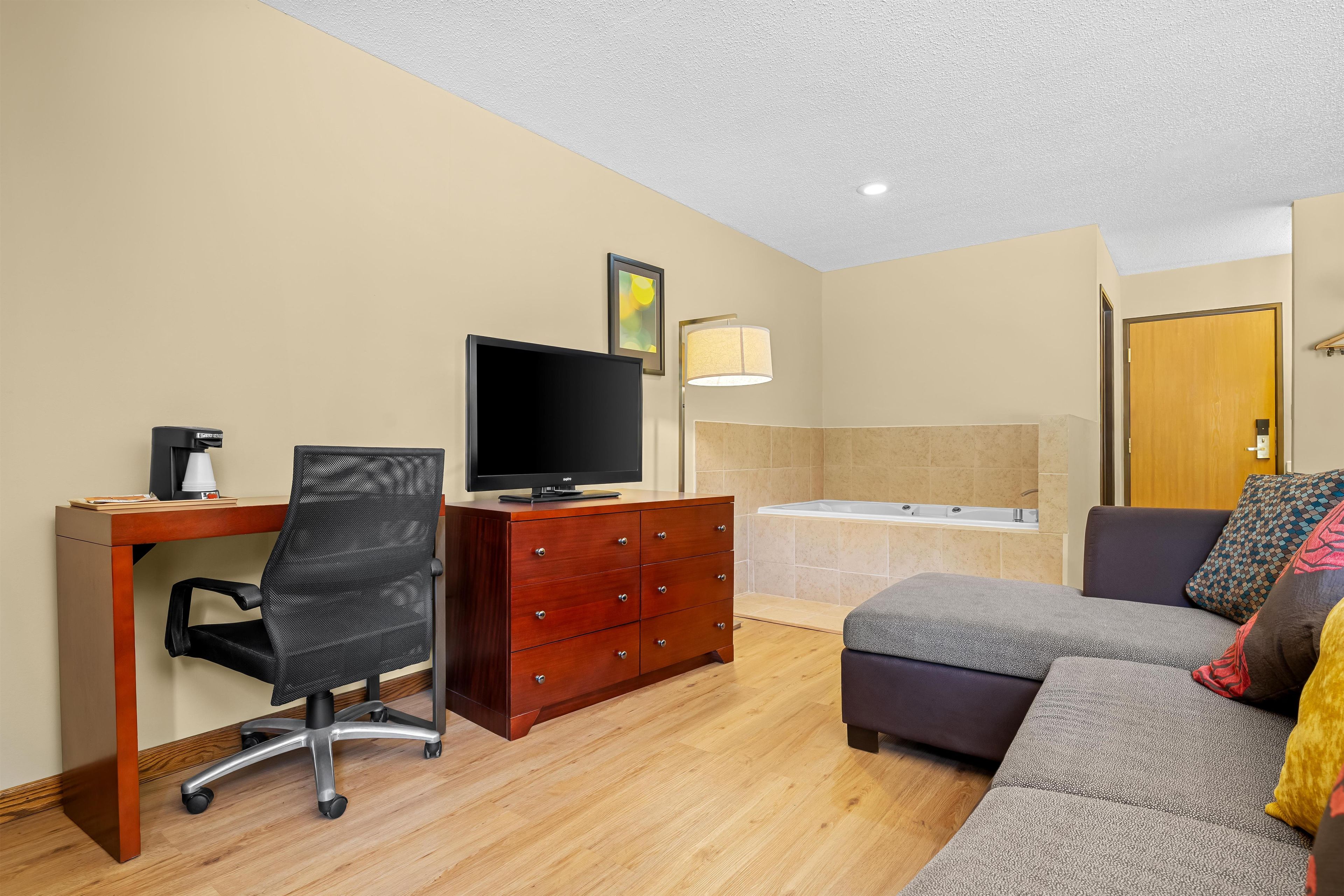A hotel room with a wooden desk, a black office chair, a flat-screen TV, a wooden dresser, a sofa, and a bathtub.