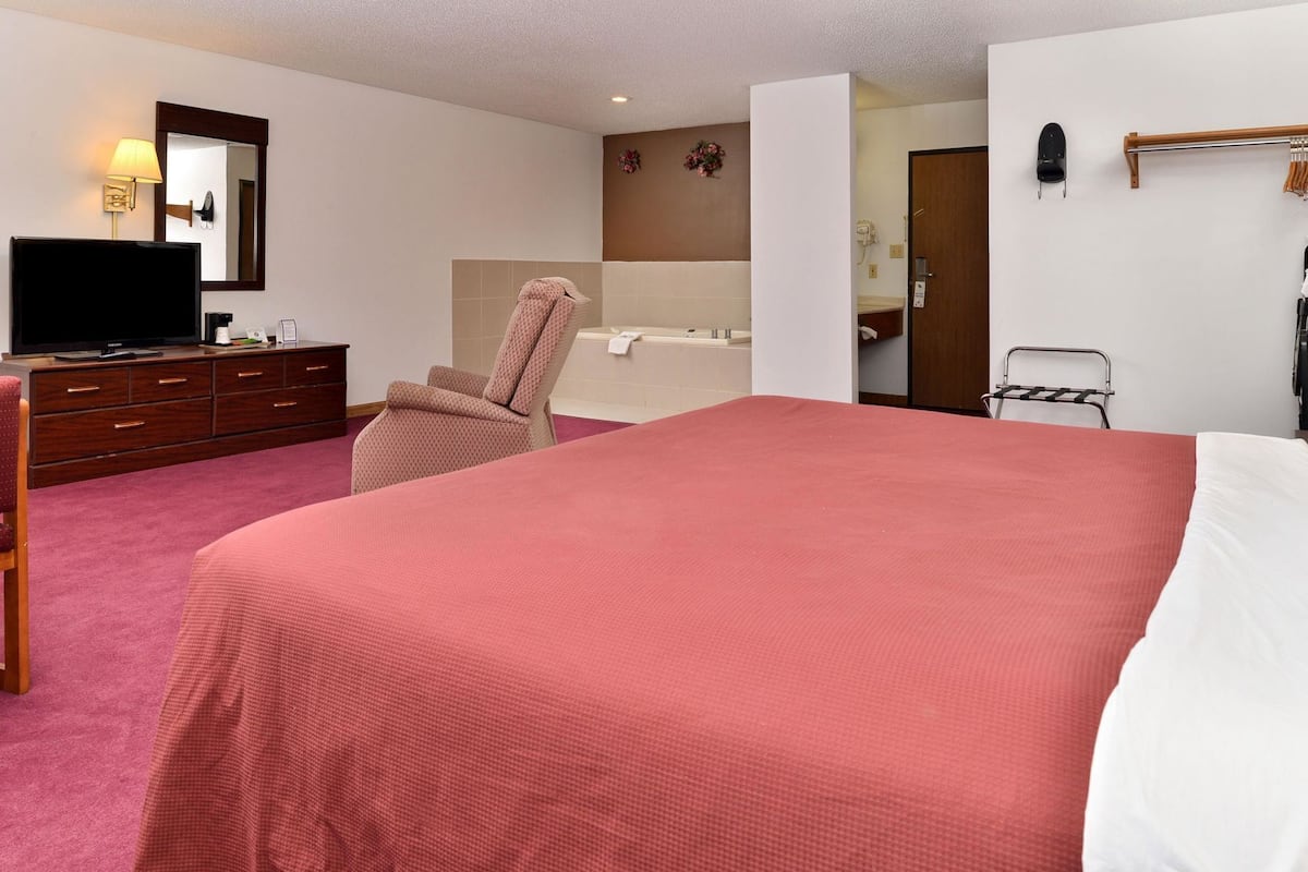Une chambre d’hôtel avec un grand lit, une chaise, une télévision, une commode, une salle de bain et un porte-manteau.