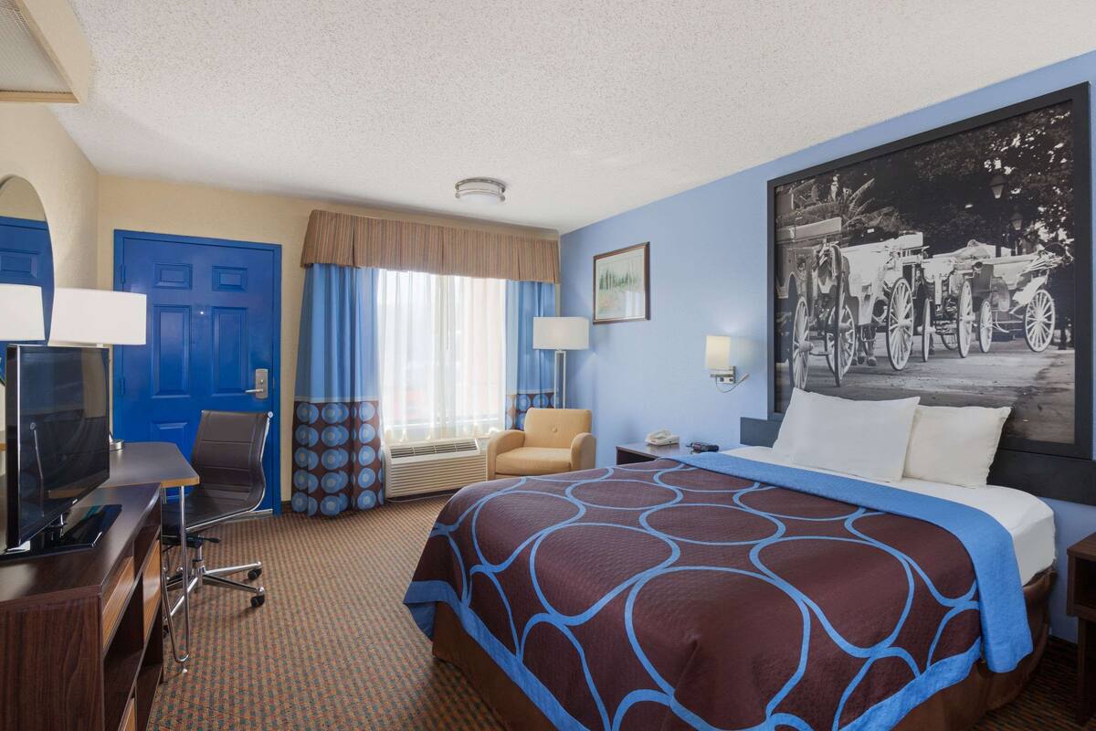A hotel room with a large bed, a desk with a computer, a chair, a window with curtains, a wall-mounted TV, and a large framed picture.