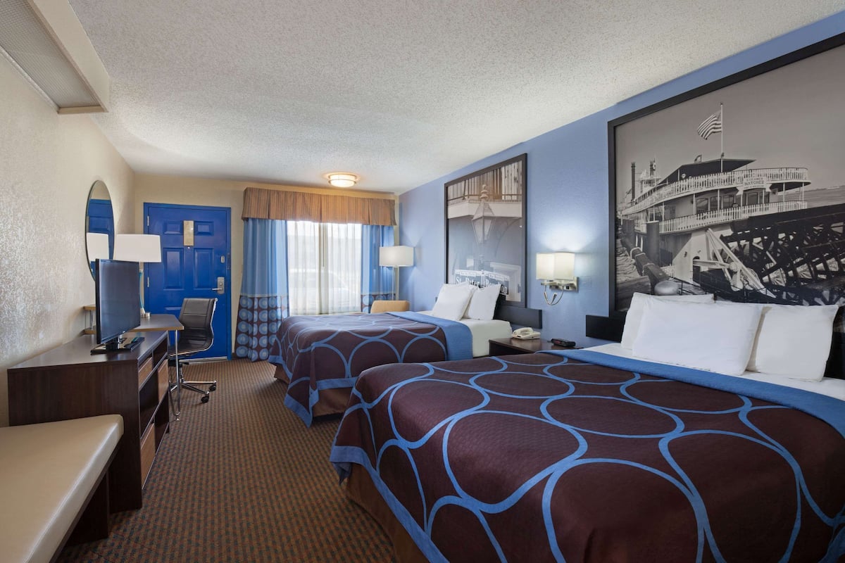 A hotel room with two beds, a desk, a chair, a TV, and a large framed picture on the wall.