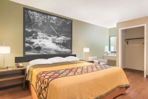 A hotel room with a large bed, a nightstand with a lamp, a desk with a phone, a bathroom with a mirror, and a door leading to another room. - Super 8 by Wyndham Bryant Little Rock Area (Bryant)