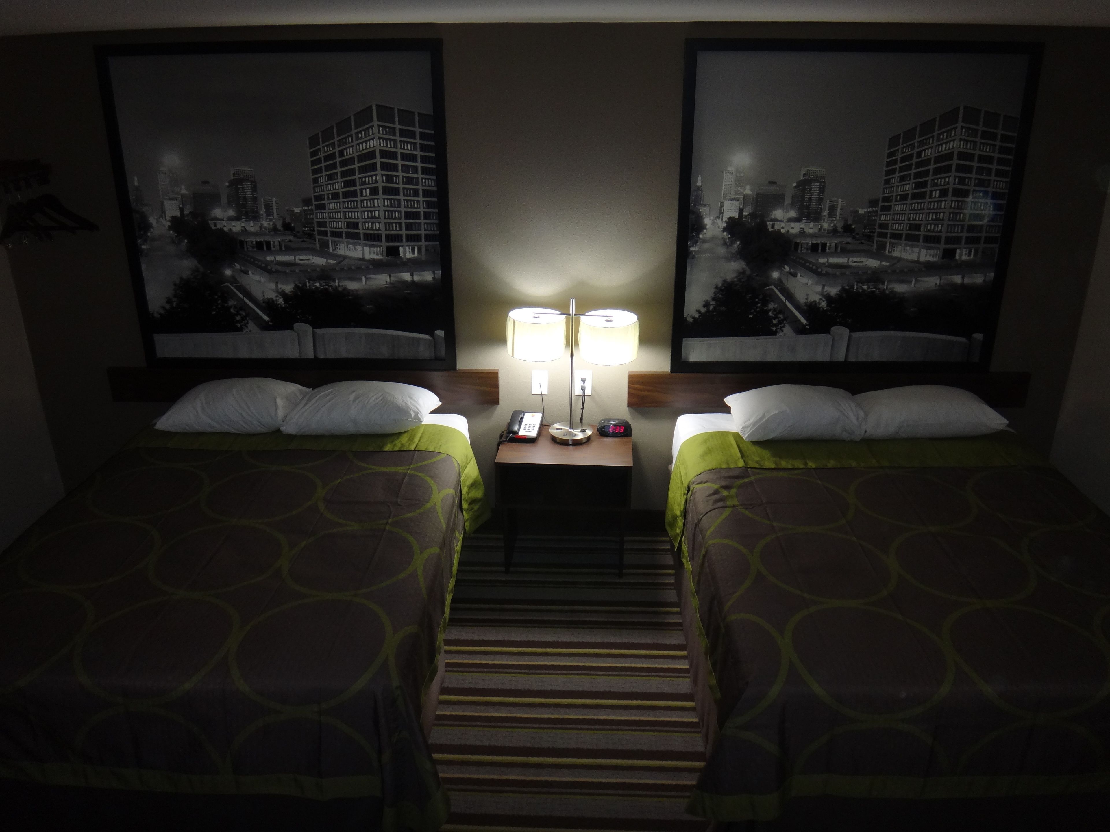 A hotel room with two beds, a nightstand with a lamp, and a framed cityscape picture on the wall.