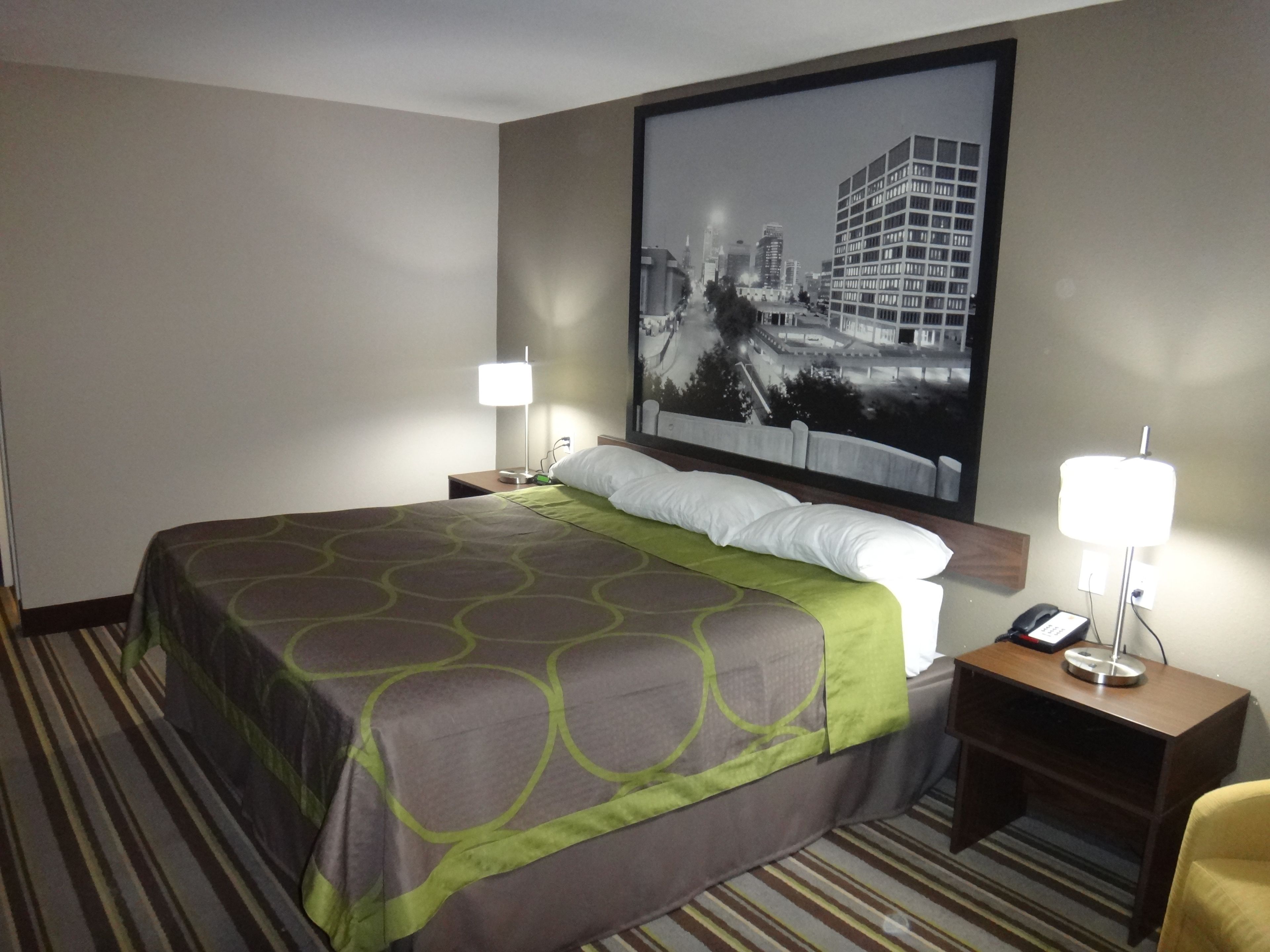 Quarto de hotel com uma cama grande, duas mesas de cabeceira, um criado-mudo, um telefone e uma pintura com vista para a cidade na parede.