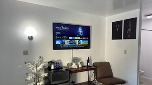 Una sala de estar moderna con un televisor de pantalla plana montado en la pared reproduciendo una película, un refrigerador pequeño, un escritorio de madera, una silla marrón y una planta de orquídea decorativa.