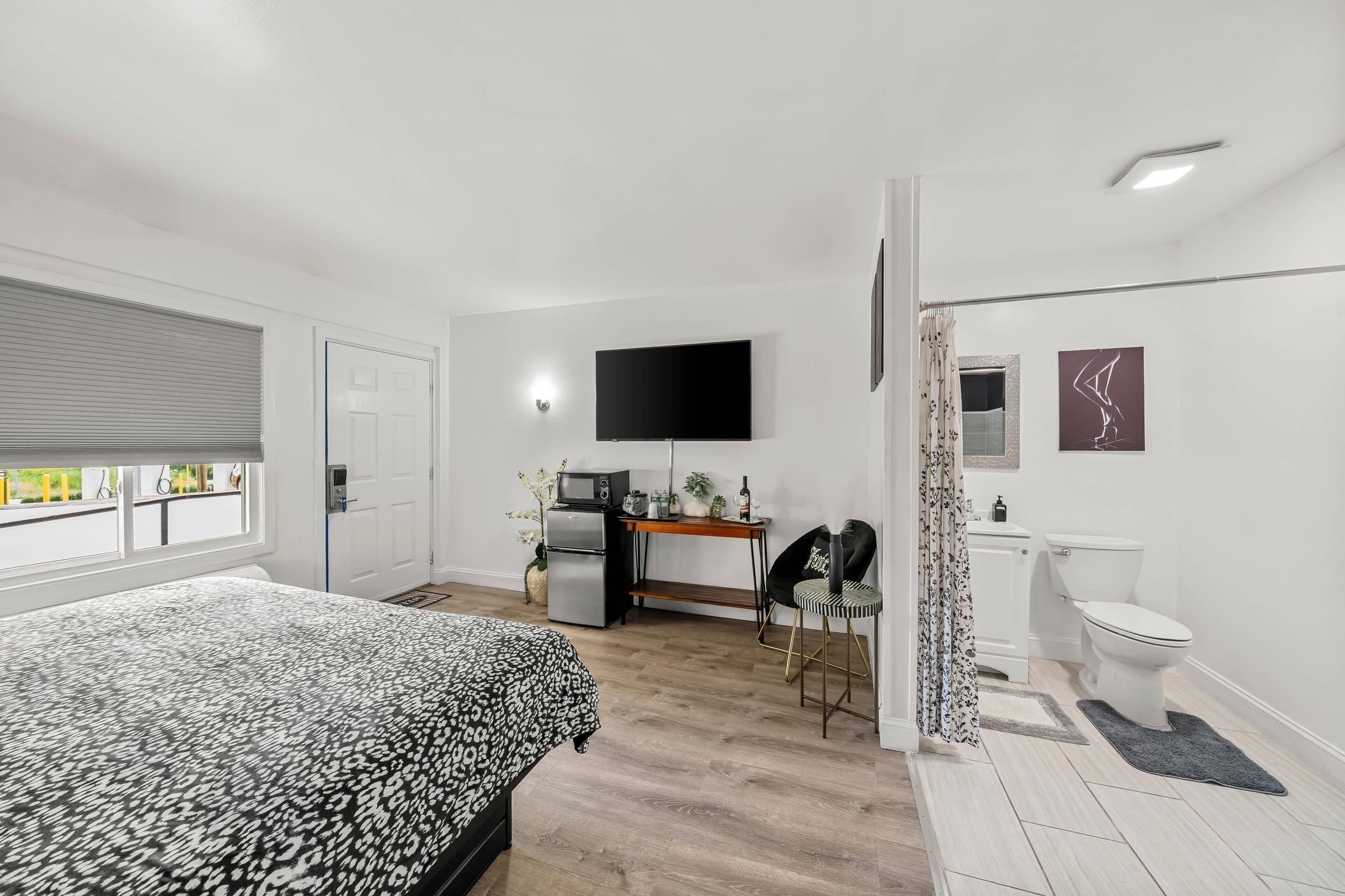 A modern bedroom with a bed, a desk with a flat-screen TV, a refrigerator, and a chair.