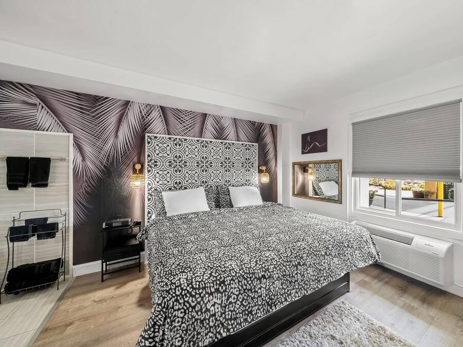 A bedroom with a patterned bedspread, a nightstand with a lamp, a wall-mounted mirror, and a window with a view of the outside.