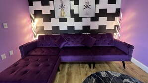 Un sofá seccional morado con diseño acolchado, un adorno de pared geométrico blanco y negro, y obras de arte enmarcadas arriba.