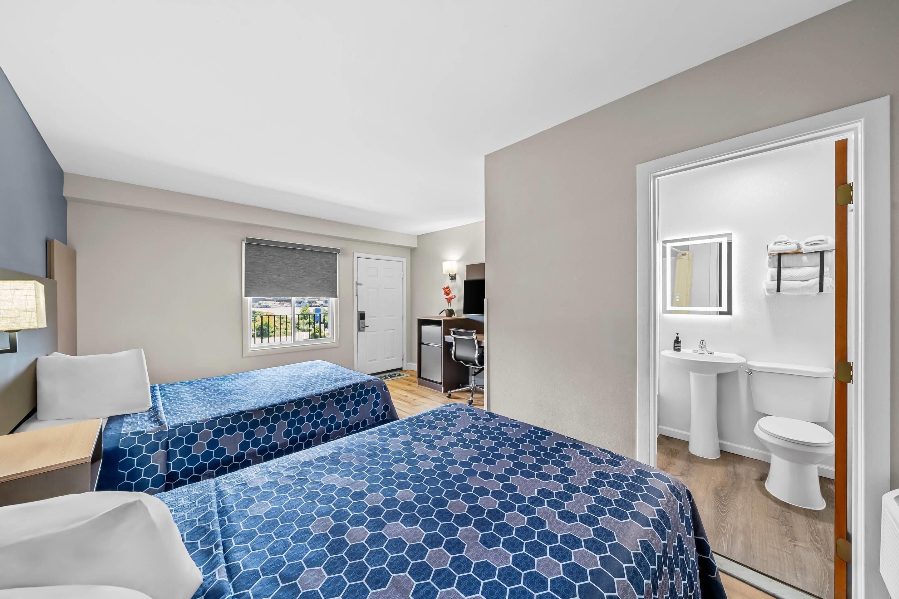 A hotel room with two beds, a desk, a chair, a window with a view, a kitchenette, and a bathroom.