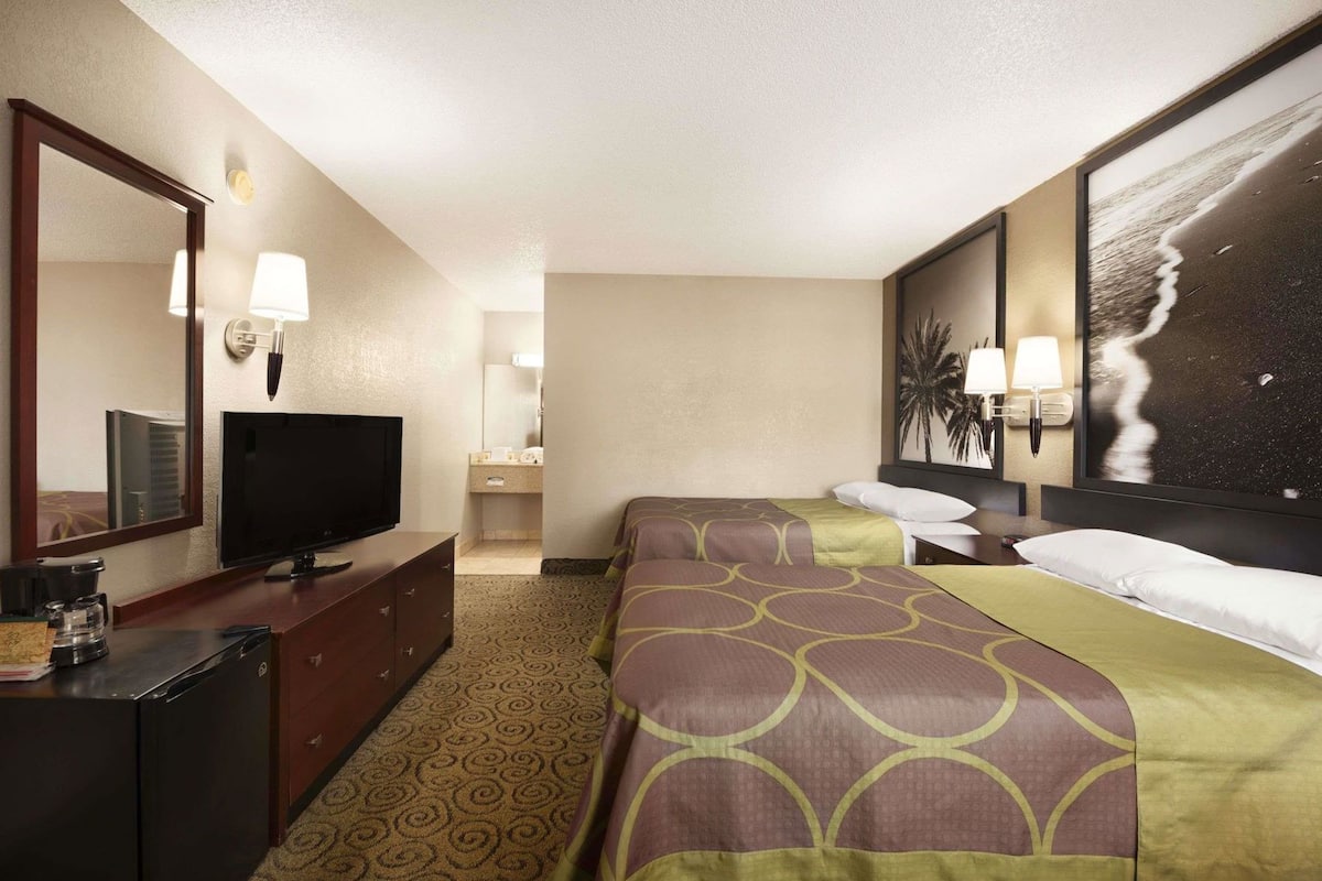 Habitación de hotel con televisión, escritorio con cafetera, dos camas con ropa de cama estampada, un cuadro grande en la pared y un baño visible al fondo.
