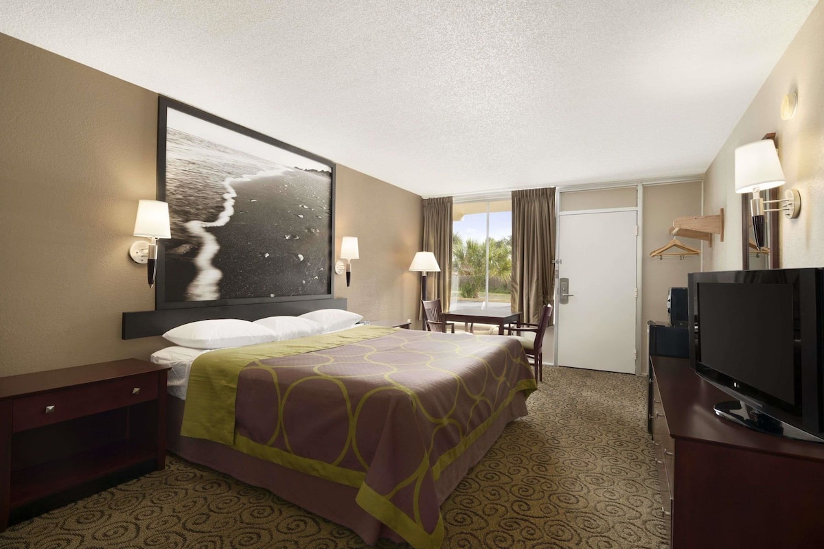 Habitación de hotel con cama, televisión, escritorio y silla. Cuenta con un ventanal con cortinas y una puerta que da al exterior.